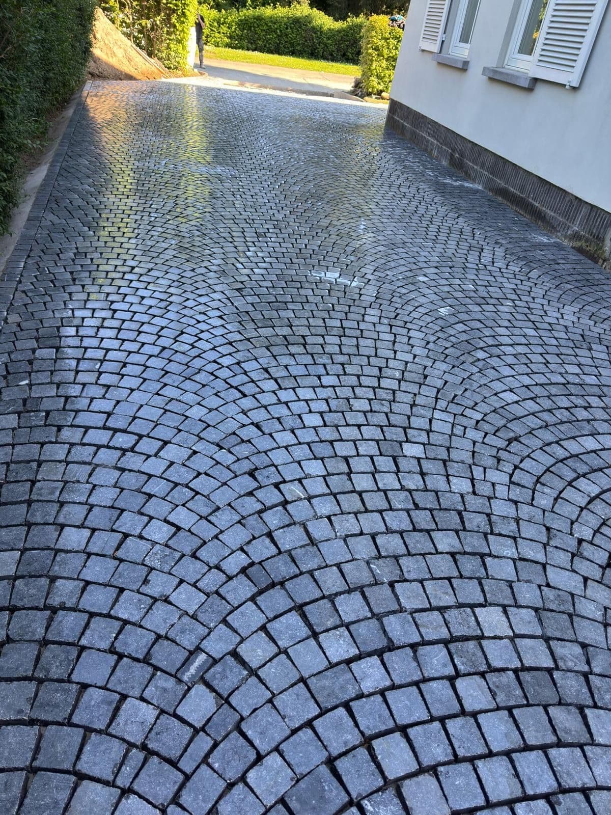 Allée en pavés posés en arcs, le long d'une maison blanche, bordée de haies